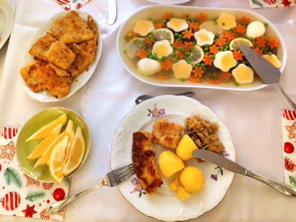 A Polish Christmas Eve table photographed from above on a white cloth with Christmas-patterned napkins: an oval dish of fish fillets fried in breadcrumbs, a long platter of fish set in clear jelly and decorated with lemon slices, green peas, carrot cut into stars and flowers of egg white, a green dish of lemon wedges, and a flowered plate holding a fried fillet, boiled potatoes and stewed sauerkraut with mushrooms, with a knife and fork laid across it.