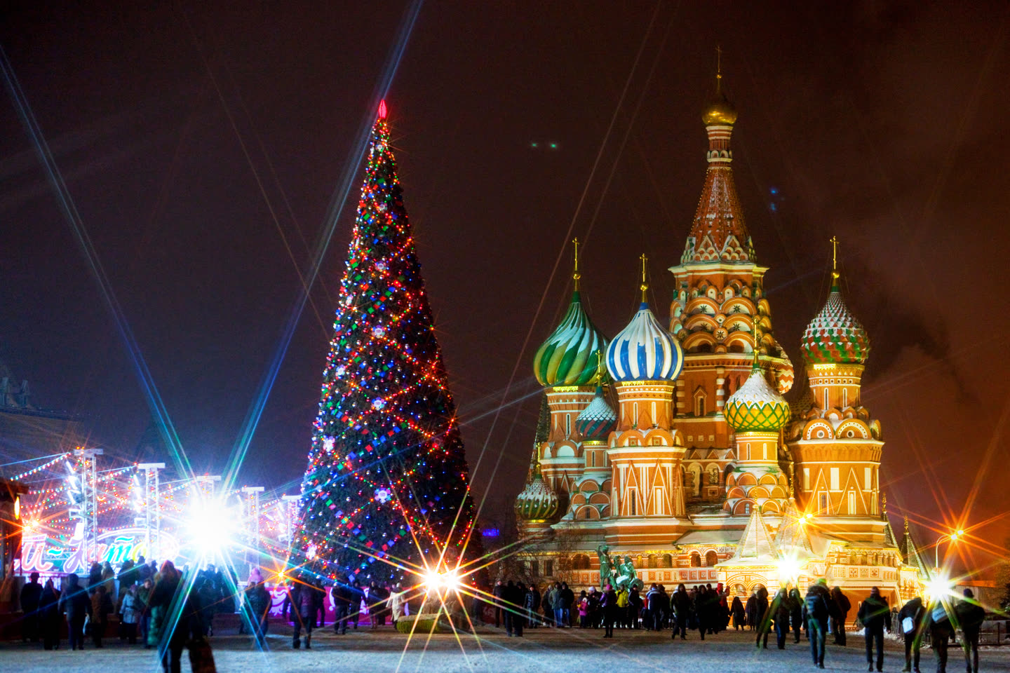 Red Square at night under floodlights: a tall conifer strung with coloured lights and topped by a red star stands at the left, Saint Basil's Cathedral rises at the right with its patterned onion domes and a gold cupola and cross above the central tent spire, and lit market stalls and a crowd fill the snowy square below.
