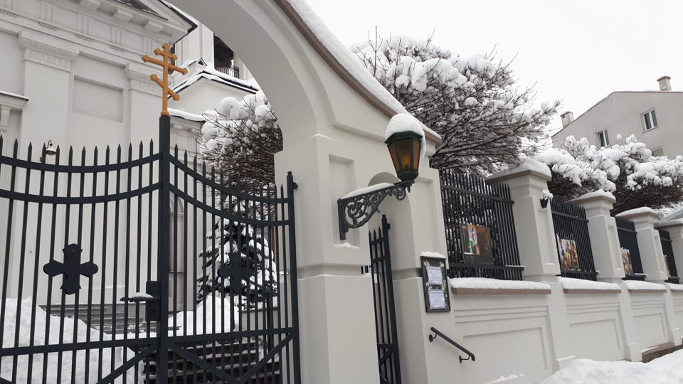 The gate of the Orthodox cathedral of Saint Nicholas in Białystok under deep snow: a whitewashed arched gateway topped with a gold three-barred Orthodox cross, black iron gates standing open onto swept steps, a wrought-iron lantern capped with snow, and icons in frames mounted along the white boundary wall. Clipped trees behind the wall are bowed under snow and a block of flats stands beyond.