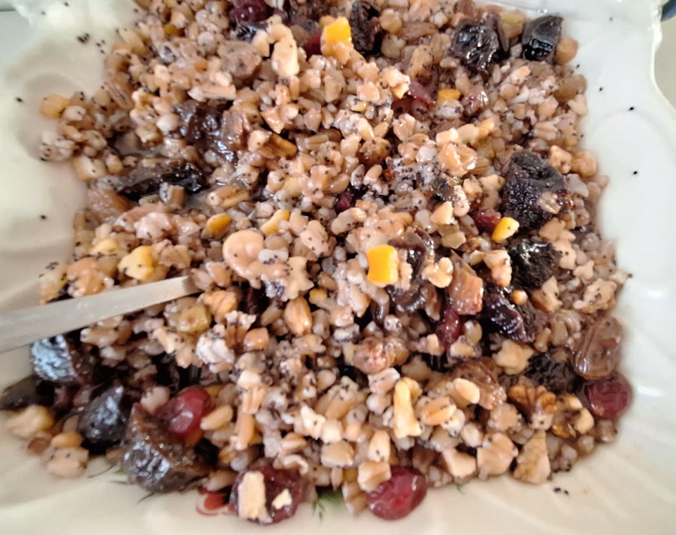 A dish of kutia seen from above: boiled wheat berries glossy with honey, scattered all through with poppy seed and chopped walnuts, with raisins, dried cranberries and pieces of dark dried fruit among them, and the handle of a spoon resting in at the left.