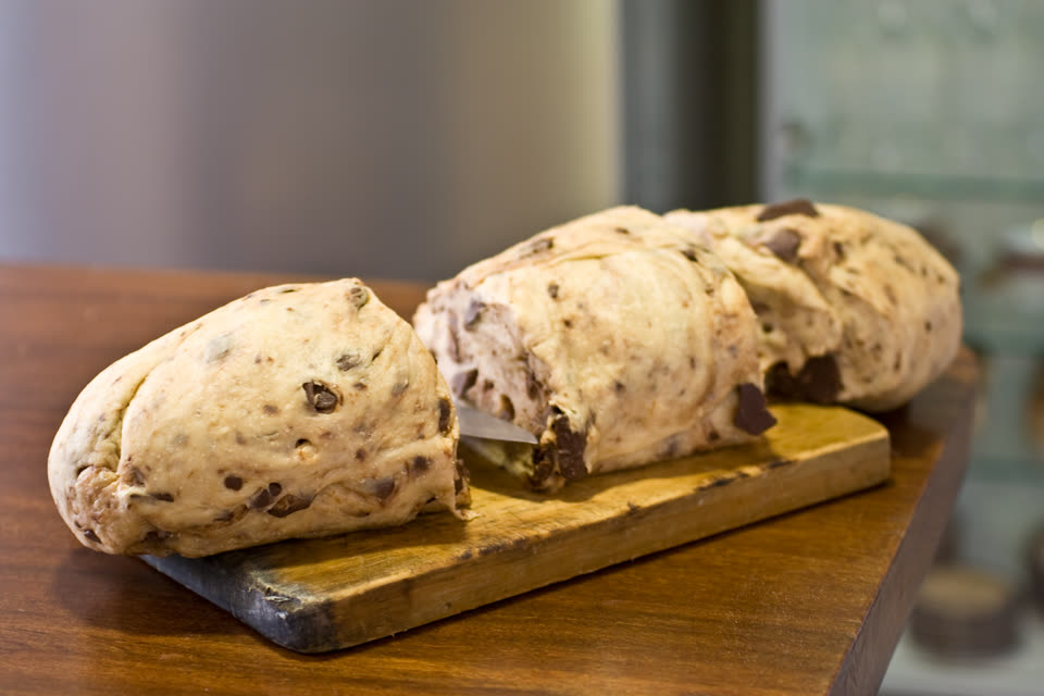Three loaves of raw chocotone dough resting on a worn wooden board on a dark wooden table, photographed from just above the table edge with the background thrown out of focus. The dough is pale cream and slack, slumping gently under its own weight, and is packed with irregular chunks of dark chocolate that break the surface all over — some pressed flat, some standing proud at the cut ends. The nearest loaf is whole and rounded; behind it two more are pushed together, with a small slip of white paper tucked between them. Nothing is baked yet and there is no tin, no icing and no wrapper. No people are in the picture.