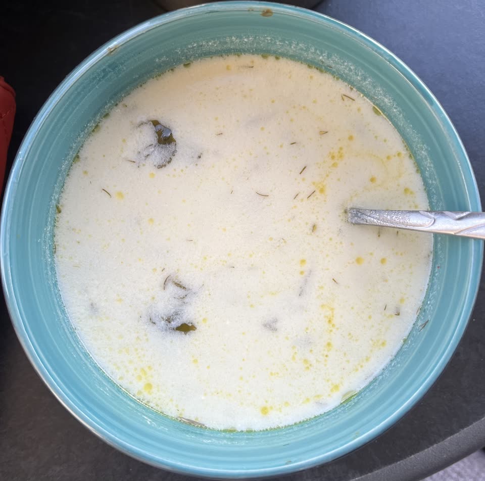 A wide turquoise bowl of spas, the Armenian yogurt soup, filled nearly to the brim and pale as cream, with dried herbs and dark green leaves showing through the surface and beads of yellow butter floating on top; a spoon rests in the bowl.