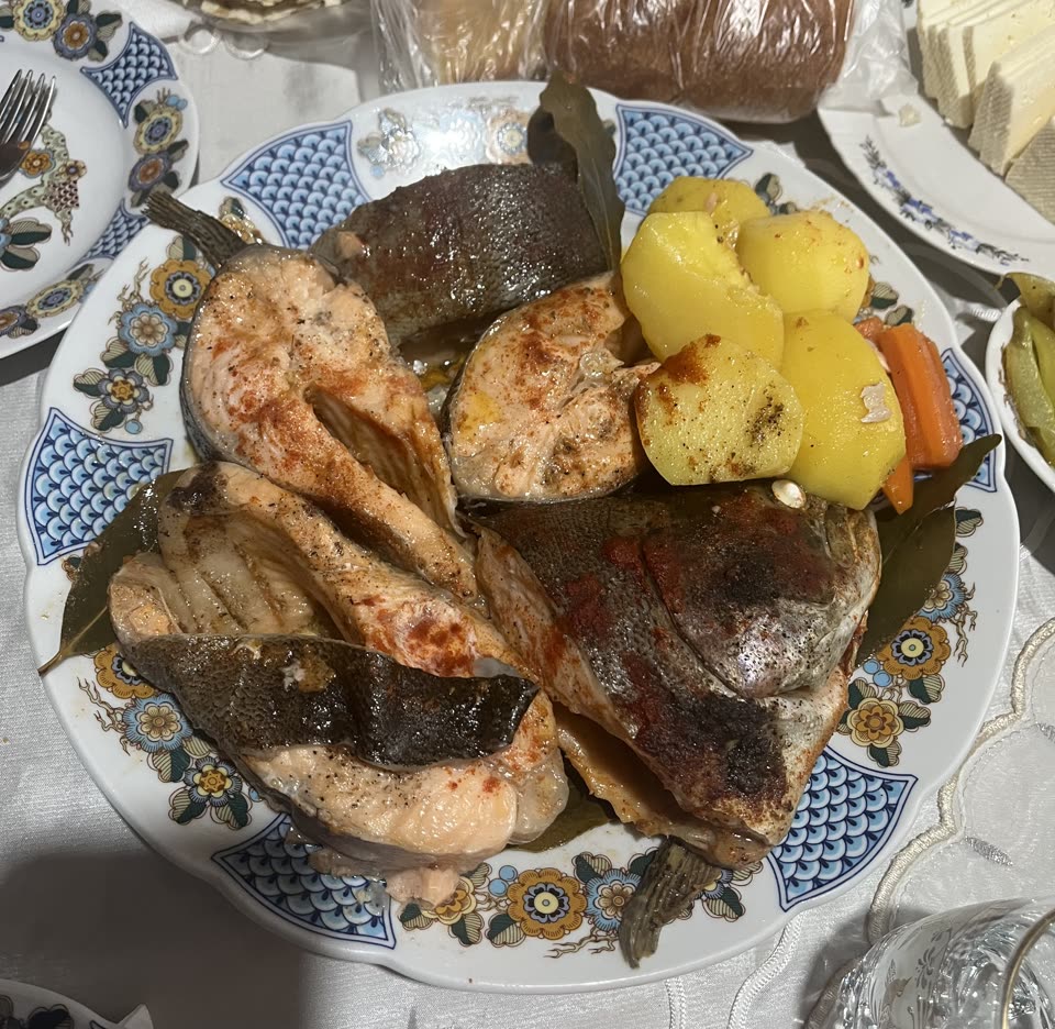A blue and white patterned platter of stewed fish on a laid table: thick cut steaks of pink trout dusted with paprika, a whole fish head among them, bay leaves tucked between, and boiled potatoes and carrot heaped to one side. Cheese, pickles and bread stand on the cloth around it.