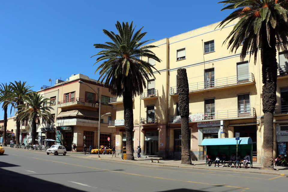 Harnet Avenue in Asmara in hard midday sun, seen from across an empty tarmac road marked with a yellow centre line. Tall date palms with thick scaly trunks stand along the near pavement and throw sharp shadows over it. Behind them is a pale yellow modernist block of three storeys, flat-fronted, with small railed balconies, square windows and a plain parapet; an older ochre building with a deep shaded verandah adjoins it on the left. Shops fill the ground floor, one signed SENEBI INTERNATIONAL TRAVEL AGENCY and another lettered Cafe. A green bus shelter with a curved roof stands at the right with about eight people sitting along its bench. A white van and a yellow taxi are stopped further up the street, bicycles are parked at the far left, and the sky above the roofline is deep and cloudless blue.