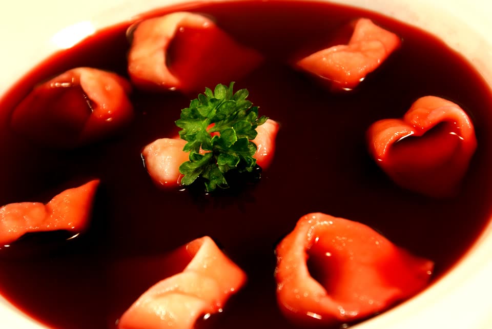 A wide white bowl of barszcz seen from above: a clear, deep crimson beetroot broth with seven uszka floating in it — small pale dumplings pinched into rings — and a sprig of curly parsley in the middle.