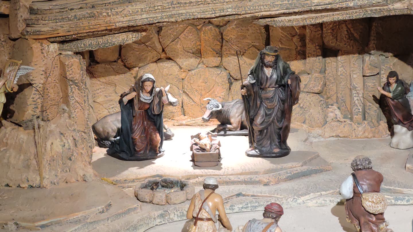 The Nativity at the heart of a Spanish belen, in painted clay figures a few inches high. Mary kneels on the left in a dark blue cloak and Joseph stands on the right leaning on a staff, both looking down at a low wooden manger where the child lies on straw in the one patch of bright light in the scene. An ox lies behind the manger with its head turned outward and a donkey stands in shadow at Mary's shoulder. The stable is built up out of rough cork bark and moulded rock with a heavy timber beam across the top of the frame. Along the front edge, below the floor of the cave, three shepherds climb towards it with blanket rolls and water gourds slung across their backs, and the gilded wing of an angel shows at the far left.
