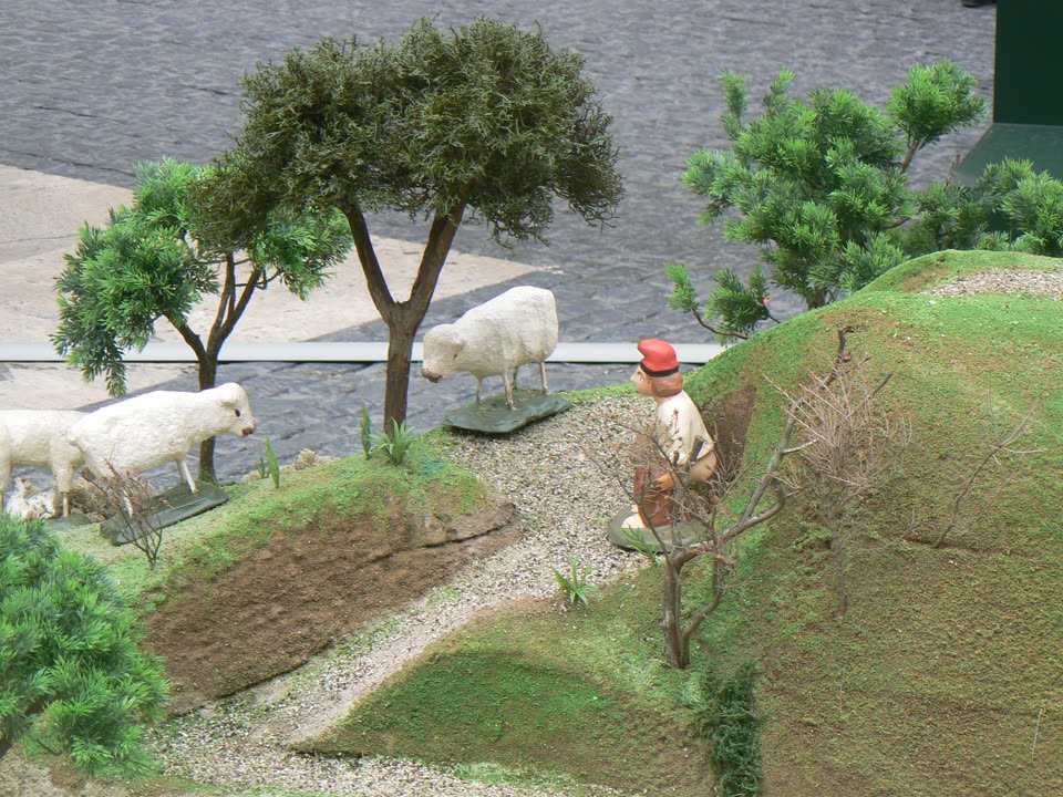 A caganer in place in a Catalan nativity scene, found rather than displayed. The figure is a small man in a red barretina and a pale shirt, squatting with his trousers down and his back half turned, tucked into a hollow on a mossy slope behind a bare twiggy bush that mostly hides him. Around him the crib is built out of real moss, gravel paths and miniature conifers, with three white model sheep grazing on the terrace above and to the left. Beyond the model landscape the photograph shows what the crib is standing on — a slab of grey paving and a dark painted edge — so the scale is unmistakable.