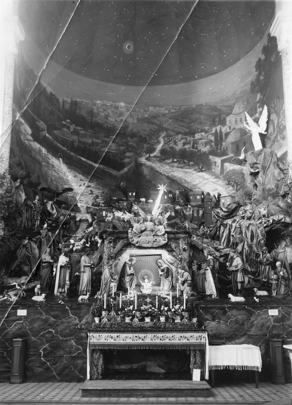 A photograph of 1933 of the Christmas crib in the Franciscan basilica at Panewniki in Upper Silesia: an enormous painted panorama of Bethlehem curving across the apse, its white houses and domes spread over hills under a starry sky, and in front of it a built foreground of rock and moss carrying life-size figures — shepherds, kings, an angel with spread wings high at the right — all turned towards the Nativity at the centre, where the Child lies lit beneath a star while Mary kneels. Candles and potted flowers stand on the altar shelf below. A crack in the glass plate runs diagonally across the picture.