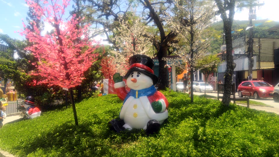 A large fibreglass snowman sitting on a bank of vivid green grass in hard midday sunshine. He wears a black top hat with a red band, a blue and yellow scarf, a red and white coat with yellow buttons and green mittens, and he has a carrot nose and black boots; one arm is raised in a wave. The grass around him is lush and freshly cut, and the sun throws sharp tree shadows across it. Behind him stand artificial blossom trees, one bright pink and one white, and beyond those are real tropical trees in full leaf, a red and white candy-cane pole, and a row of small shopfronts along an ordinary street with cars parked outside, including a red hatchback at the right. The sky above is deep blue with white cloud. A second, smaller figure in a bowler hat sits further back at the left. A few people are visible at a distance on the pavement and near the shops, and one figure stands partly cropped at the extreme left edge. Nothing in the picture is cold.