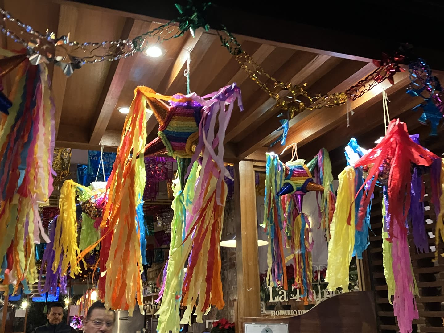 Half a dozen traditional star piñatas hanging close together from the beamed wooden ceiling of an indoor market hall, photographed looking up. Each is a ball of coloured tissue with seven cones radiating from it, and from the tip of every cone falls a long bunch of crepe paper streamers in pink, yellow, orange, lime, purple and blue, so that the whole ceiling is curtained in paper. The nearest piñata is banded in rainbow stripes with a gold cone pointing at the camera; others in red, white and blue hang behind it. Foil garlands in silver and gold are strung between the beams, and papel picado in blue and purple shows through the gaps beyond. Warm downlights are set into the ceiling. Two men stand at the bottom left of the frame, well below the piñatas and partly cut off by the edge.
