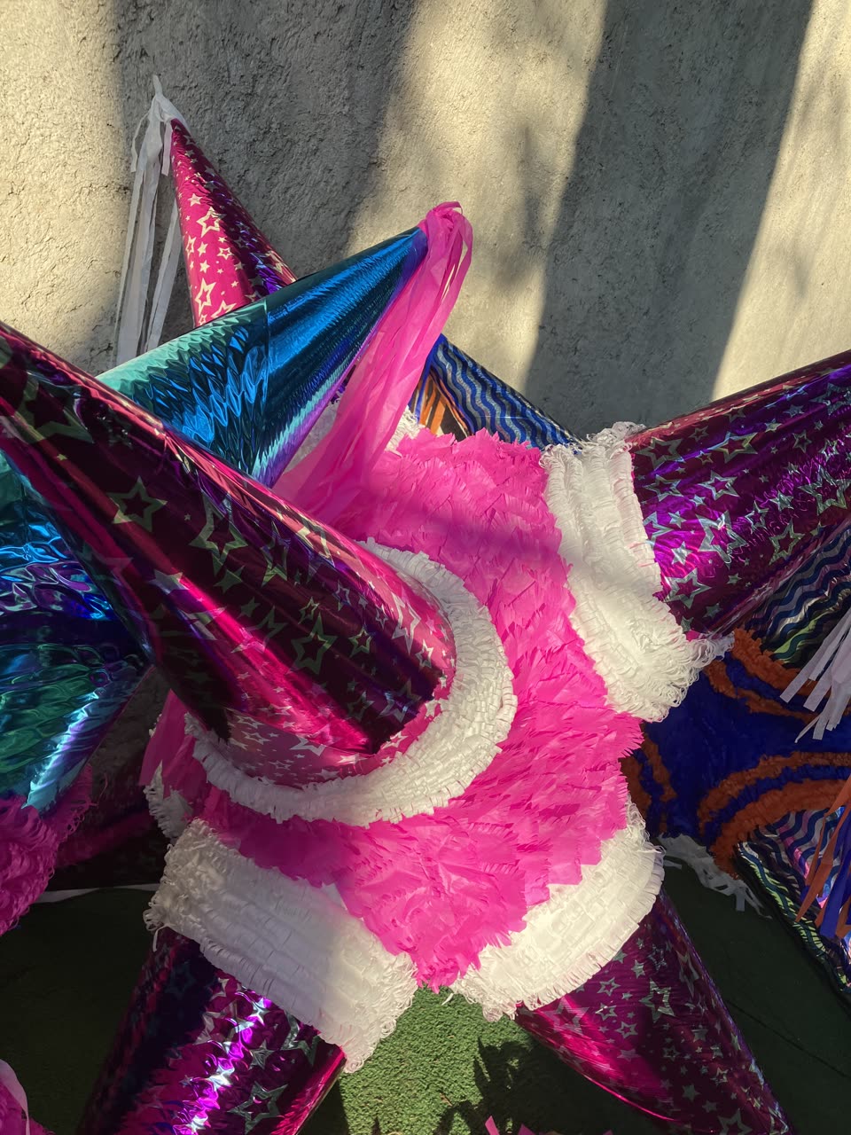 A close view of a single star piñata standing against a rough grey stucco wall in hard sunlight, filling the frame. Its cones are wrapped in metallic foil printed with stars — magenta, hot pink, electric blue and teal — and each cone is finished at the base with a fringed collar of crepe paper in bright pink and white. A second piñata in blue and orange stands behind it at the right, and a third shows at the left edge. The fringing is cut into fine strips and ruffled in overlapping rings, and loose white paper tails hang from two of the points. The ground beneath is green artificial turf and the shadow of foliage falls across the wall. No people are in the picture.