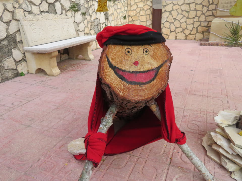 A village tio set up on a paved square, big enough to sit on. It is a thick sawn log, the end facing the camera painted with two round eyes, a small red nose and a wide grinning mouth, with a red and black barretina pulled over the crown and a red fleece blanket draped down its back like a cape. Two stripped branches hold up the front of it, with cloth wrapped around one as a sleeve, and the whole thing stands on a stone slab in front of a carved stone bench and a dry-stone wall in the warm afternoon light.
