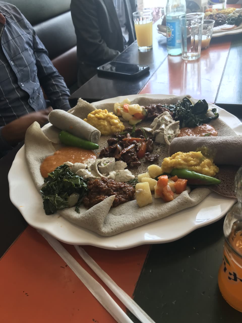 A wide white plate on a cafe table, lined with a single grey-brown injera whose surface is pocked all over with small holes. The bread is both the plate and the food: ten or so dishes are spooned directly onto it in a ring — chopped dark greens, bright yellow split peas, diced potato and carrot, a smooth orange-red sauce, a white curd-like dish scattered with herbs, a dark stew of meat with onion and tomato in the centre, and two whole green chillies. Two tightly rolled injera sit at the back like napkins. Beyond the plate: a dark table with a phone, tumblers, a bottle of mineral water and a glass of orange juice, a second platter at the top right, and two seated people visible only as a check-shirted torso and an arm.