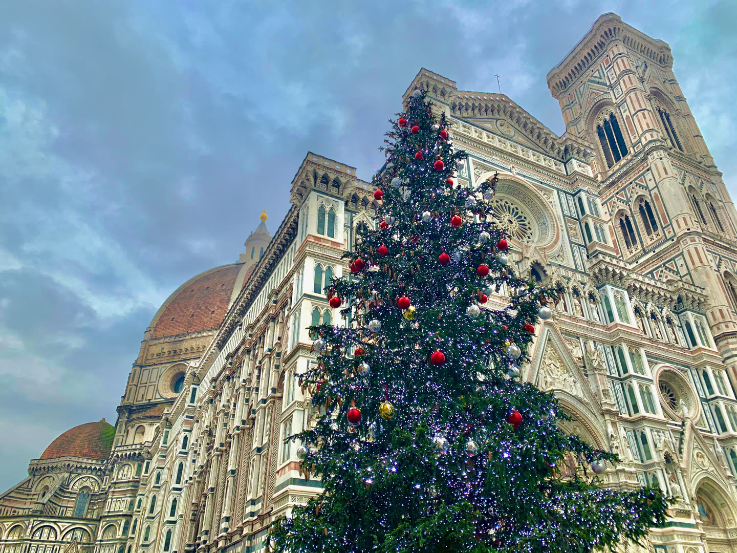The Christmas tree in the Piazza del Duomo in Florence, standing against the cathedral of Santa Maria del Fiore. The tree fills the right half of the frame, a tall dark fir strung with white lights and hung with large red and silver baubles and one gold one. Behind and above it the cathedral's flank rises in bands of green, white and pink marble: the arcaded gallery, a carved rose window, the pointed gables of the façade, and at the far right the lower stages of Giotto's campanile in the same three marbles. At the left, over the roofline, Brunelleschi's brick dome shows its red tiles, white ribs and the lantern above, with the smaller green-and-white dome of the Baptistery below it. The sky is overcast in grey and pale blue.