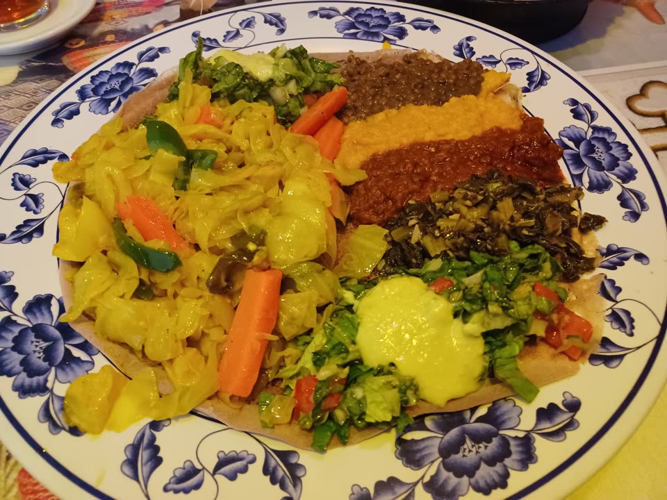 A single round of injera laid over a blue-and-white floral china plate, serving as the plate itself, with the food spooned directly onto it and no bowls anywhere. The bread is pale greyish-brown and faintly porous at the edges where it shows. Across the left half is a heap of yellow turmeric-stewed cabbage with potato chunks, strips of green pepper and whole batons of carrot. The right half carries five smaller mounds pressed up against one another: dark green-brown lentils at the top, a smooth orange puree below them, a dark red-brown stew below that, then chopped dark greens, and at the bottom a fresh salad of shredded lettuce and diced tomato with a pale yellow-green dressing poured over it. There is no meat, egg or cheese on the plate. A white dish and a patterned cloth are out of focus beyond the rim.
