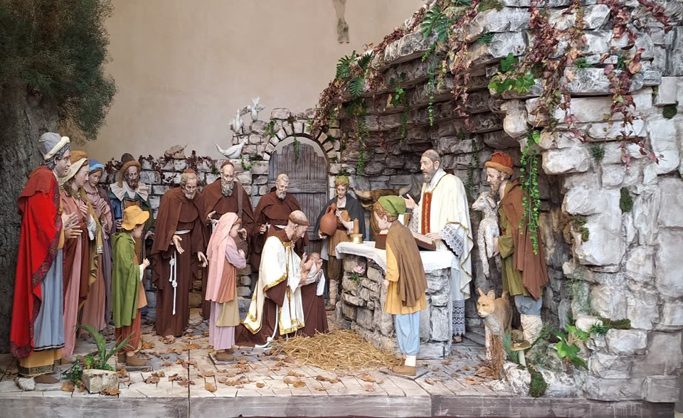 A modelled nativity scene that depicts the first one: not Bethlehem but Greccio. Life-size painted figures stand in a stone-walled recess hung with ivy and vine. At the centre a friar in a brown Franciscan habit and a white and gold stole holds the swaddled child above a bed of loose straw on the floor. To the right a priest in a red and white chasuble stands at a rough stone altar with a chalice on it, saying Mass over the manger. Villagers fill the left half — a woman in red, children, a boy in green — and more friars in brown habits stand among them. A shepherd at the right carries a lamb across his shoulders, an ox looks out from behind the altar, a dog sits in the foreground and a dove is fixed to the wall above.