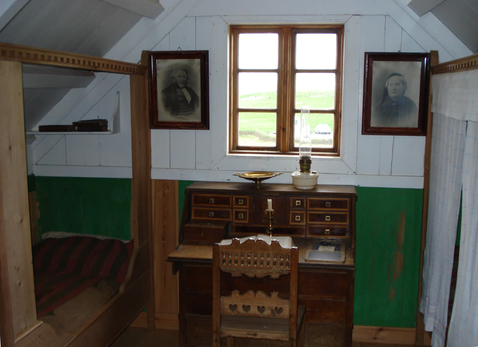 A room in the turf farm at Glaumbaer: a wooden writing bureau under a small window with its flap down and an open book on it, a carved chair with heart cut-outs, an oil lamp and a brass candlestick, framed portraits either side of the window, and a box bed with a striped blanket in the alcove to the left.