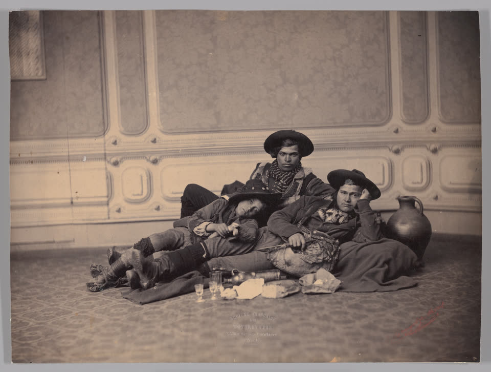 A nineteenth-century studio photograph of three pifferari lying on the floor of a Paris photographic studio, against a painted backdrop of panelled wall. All three wear broad-brimmed felt hats, short jackets and the cross-gartered leggings of the central Italian mountains, with rough hide shoes. The man at the centre is propped on one elbow, half asleep; the one behind him looks straight at the camera; the third, at the right, rests his head on his hand. Their bagpipe lies across the middle man's lap with the skin bag slack. Spread on the floor in front of them are a terracotta jug, two small stemmed glasses, a loaf torn open and a wedge of cheese on paper. The mount is printed SOCIETE FRANCAISE DE PHOTOGRAPHIE, PARIS.
