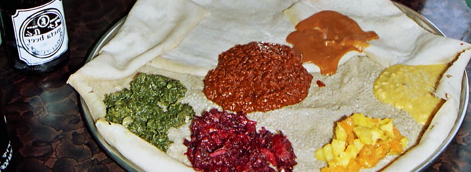 A round metal tray lined with injera, the pale spongy flatbread with its honeycomb of holes, its edge folded loosely over the rim. Six mounds of stew are spooned straight onto the bread: chopped dark greens at the left, a glossy dark-red lentil stew in the middle, a smooth caramel-brown pool of shiro above it, pale yellow split peas at the right, diced potato and carrot below them, and shredded crimson beetroot at the front. There is no meat, no egg and no cheese anywhere on the tray. A dark beer bottle stands at the left edge of the frame.
