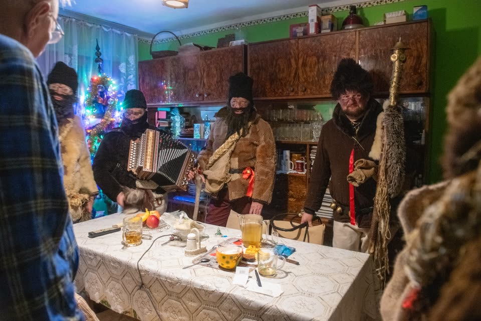 Carollers standing inside a kitchen they have been let into: a green wall, a dark veneered wall unit from the 1970s, a lit Christmas tree, and a table under a lace cloth laid with tea in glasses, apples and a jug. Three men in sheepskin coats and tall black lambskin hats, their faces behind dark balaclavas and false beards; one works a diatonic accordion, another holds a staff topped with brass. The householders stand at the edge of the frame watching.