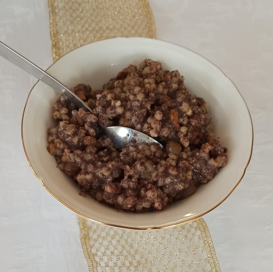 A gold-rimmed white bowl of kutia on a white damask cloth with a gold organza runner under it: boiled wheat grains darkened all through with ground poppy seed, with pieces of walnut and almond showing among them, and a spoon resting in the bowl.