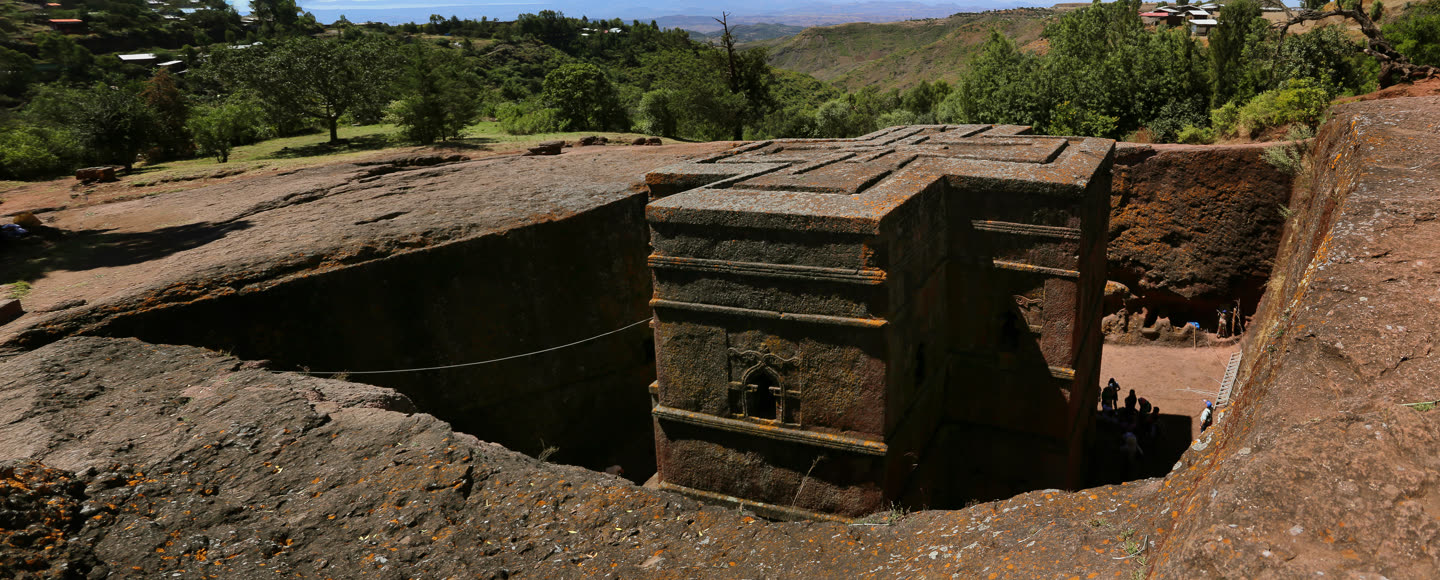 Bete Giyorgis at Lalibela seen from the rock shelf above it. The church stands in a pit quarried out of the hillside, its walls free on every side and its flat roof carved with a nested Greek cross, level with the ground the viewer is standing on. Three stepped string courses run round the walls; a tall keyhole window with a carved frame sits in the near face, and the stone is dark grey-brown and blotched with orange lichen. A trench floor of bare red earth runs round the base. At the right a cut passage leads down into the trench, where a scattering of small distant figures stands by a white ladder. Beyond the rim: green trees, a few village buildings on the skyline at the left, and the Ethiopian highlands falling away into a hazy valley.