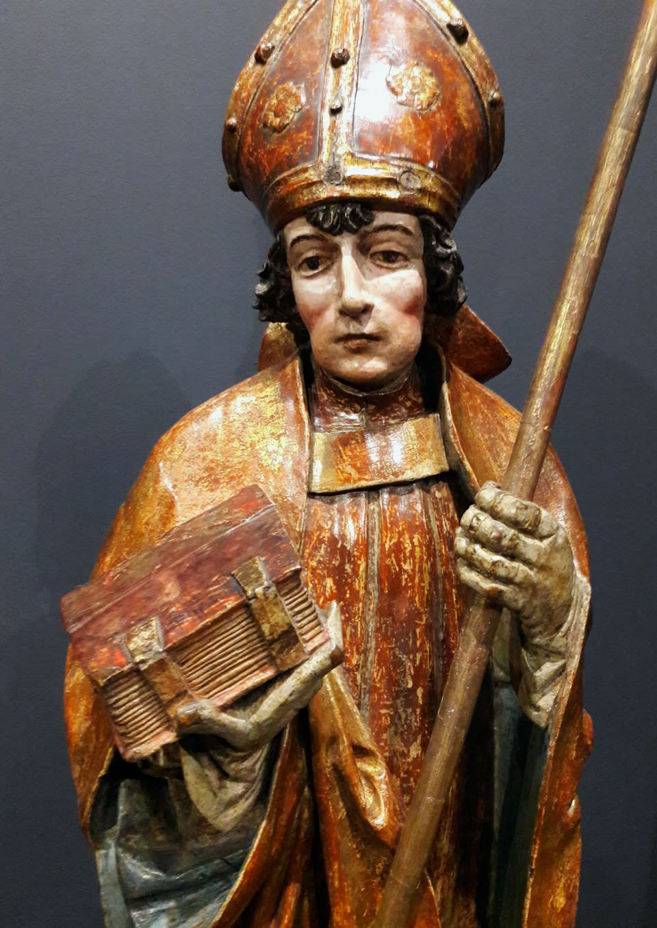 A late-Gothic carved and painted wooden figure of Saint Nicholas from Lesser Poland, photographed against a grey museum wall: a young beardless bishop in a tall red and gold mitre set with stones, dark curling hair beneath it, a red-brown cope over a gold-edged alb. He holds a thick clasped book flat on his left forearm and grips a plain wooden staff in his right hand; the paint is worn back to the gesso in places.