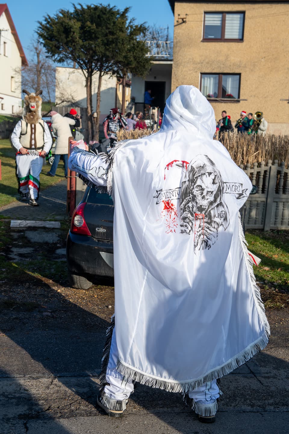 A figure in a long white hooded cape walking away along a village street, the cape printed across the back with a hooded skull holding an hourglass and the words MEMENTO MORI on a scroll, with painted blood running down from it; the white trousers and sleeves are fringed in silver. Ahead walks another costumed man in a horned animal mask and fleece, and beyond them a brass band in red hats and a figure in a printed skeleton suit stand at a garden fence.