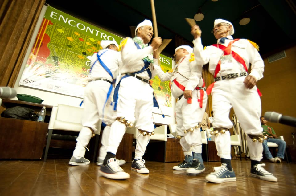 Four older men performing as a Folia de Reis company, photographed from low down on a polished wooden stage so that the camera looks up at them. All four wear white shirts and white trousers with the cuffs turned up, white caps, and broad satin sashes worn diagonally across the chest — blue on the man at the left, red and yellow on the others. Heavy black belts, bright neckerchiefs and strings of large brass bells strapped below each knee complete the costume; on their feet, incongruously, are canvas basketball boots. Two of them hold up flat wooden clappers and one grips a tall pole. Behind them hangs a large green printed banner reading ENCONTRO DE SABERES, and beyond it are stacking chairs, a table with a water bottle, and a few seated people watching from the right-hand edge of the frame. The faces of at least two of the performers are sharp and fully visible.