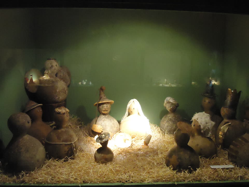 A nativity scene made entirely out of dried gourds, lit from within and displayed in a glass case against a pale green back wall. Every figure is a bottle gourd stood on its rounded base, with small bead eyes set into the shell and little else by way of a face. At the centre a tall pale gourd wears a long white veil and is lit from inside so that it glows; beside it a darker gourd in a wide straw hat holds a staff and a cupped yellow hand. Between them, low in the straw, a small split gourd stands in for the manger. The surrounding figures wear improvised headgear — a straw brim, a wrapped turban, a tin crown, a black top hat, a wisp of white wool for a beard — and three more gourd groups sit on a shelf behind. The whole arrangement is bedded in loose yellow straw that fills the bottom third of the frame. No people are in the picture.