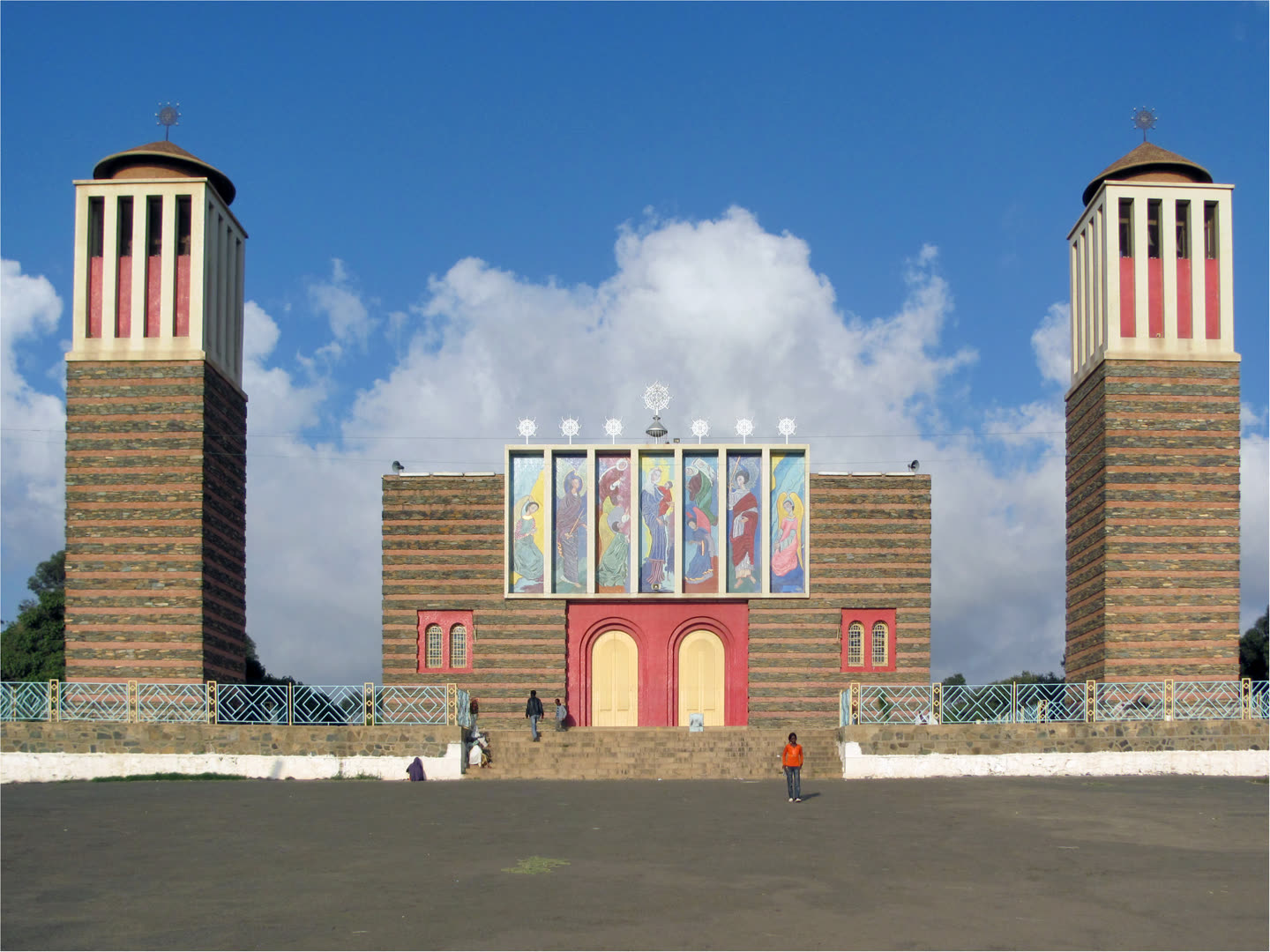 Enda Mariam Cathedral in Asmara seen head-on across an empty earth forecourt. Two tall square towers stand at either side, built in alternating courses of grey-green rubble stone and narrow red brick, each capped by a white colonnaded belfry with red panels between its columns, a low brown dome, and an openwork Geez cross on top. Between them the lower facade is banded in the same stone, with a red-painted porch and two arched cream doors at the head of a broad flight of steps. Above the doors a long panel of seven painted saints in blue, red and green robes sits in a white frame, with seven small Geez crosses in a row along its top and a larger one above the centre. A low railing patterned in blue and white diamonds runs across the front. Deep blue sky behind, with one tall white cloud rising between the towers. A few small distant figures stand on the steps and out on the forecourt.