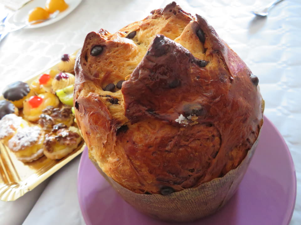 A whole panettone on a wooden board, photographed from slightly above with one thick wedge cut and leaning away from the loaf. The crust is deep brown and domed, split across the top in the characteristic cross, and the cut face shows the crumb: pale yellow, open and irregularly holed from the long sourdough rise, with dark raisins and small cubes of candied peel scattered right through it. The fluted paper case it was baked in is still around the base.