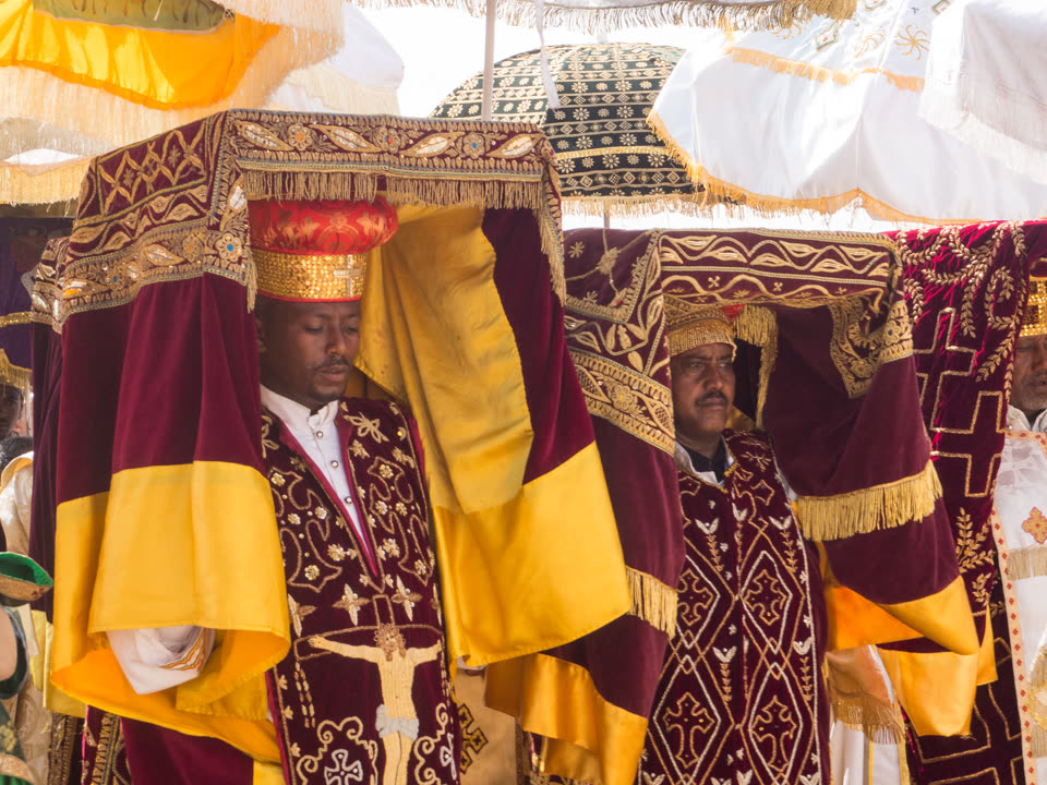 Priests carrying tabots in procession, each tabot invisible beneath the cloth that wraps it: a rectangular bundle borne flat on the head, draped in deep maroon velvet with broad bands of gold bullion embroidery and heavy gold fringe, with panels of plain yellow silk hanging below. The two nearest celebrants wear tall gold filigree crowns and maroon brocade copes worked all over with crosses, one of them carrying a long pale crucifix down the front. Above and behind, a crowd of ceremonial umbrellas fills the top of the frame — fringed, in yellow, in white, and one dark green patterned with gold rosettes. More clergy press in at both edges.