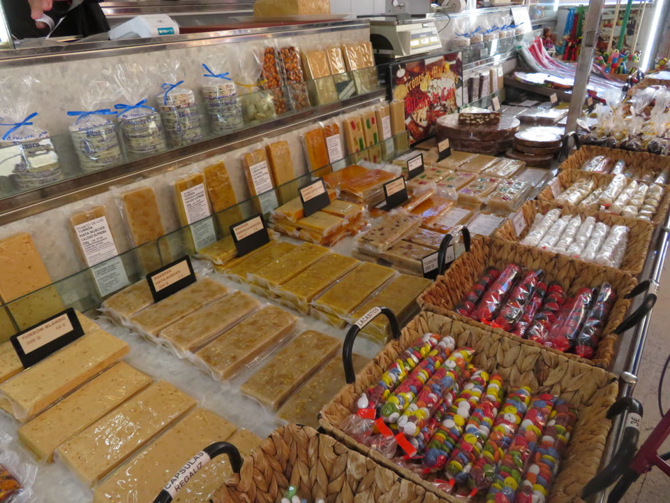 A turron stall, photographed down the length of its counter. Slabs of nougat the size of paving bricks are laid out in rows under cling film, pale cream and honey-coloured, speckled with whole almonds, each row with a small printed card propped in front of it giving the kind and the price. Behind them a glass shelf holds boxed sweets tied with blue ribbon and a set of scales; to the right, woven baskets hold wrapped sugared almonds and columns of brightly coloured jellies in cellophane. An arm and part of a torso appear at the top left corner, cropped above the shoulder, belonging to somebody working behind the counter.