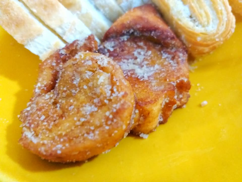 A close, slightly soft photograph of three rabanadas on a bright yellow plate. Each is a thick oval slice of bread, deep-fried to a mottled gold and reddish brown, with the crust showing darker along one edge, and all three are dusted generously with granulated cinnamon sugar that catches the light. They overlap one another in a loose row across the middle of the frame. Behind them at the top left lie several slices of plain untoasted white bread waiting to be used, and at the top right the end of a rolled, layered pastry. A scatter of loose sugar lies on the plate beside the nearest slice. No people are in the picture.