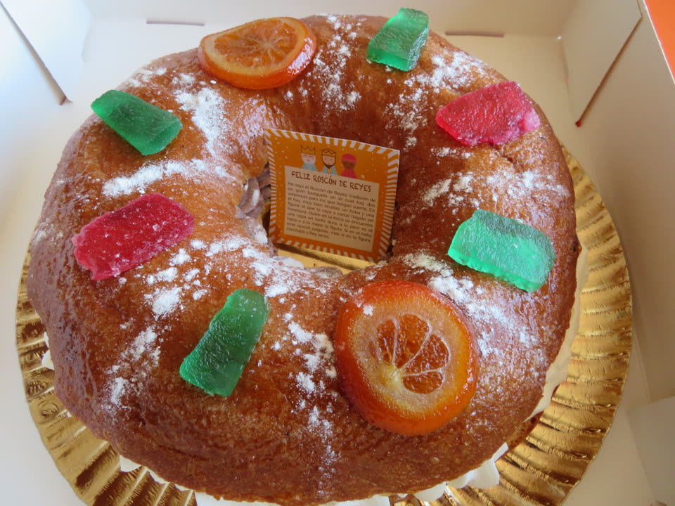 A roscon de Reyes photographed from above in the open bakery box it came in. The ring is a deep baked gold, dusted unevenly with icing sugar, and decorated with two translucent slices of candied orange and six thick pieces of candied fruit in bright emerald green and raspberry pink, set at intervals around the top. It sits on a fluted gold cake board. Standing upright in the hole in the middle is a small printed card headed Feliz Roscon de Reyes, with three cartoon kings along the top and a paragraph of Spanish explaining the two things hidden inside and what finding each of them means.