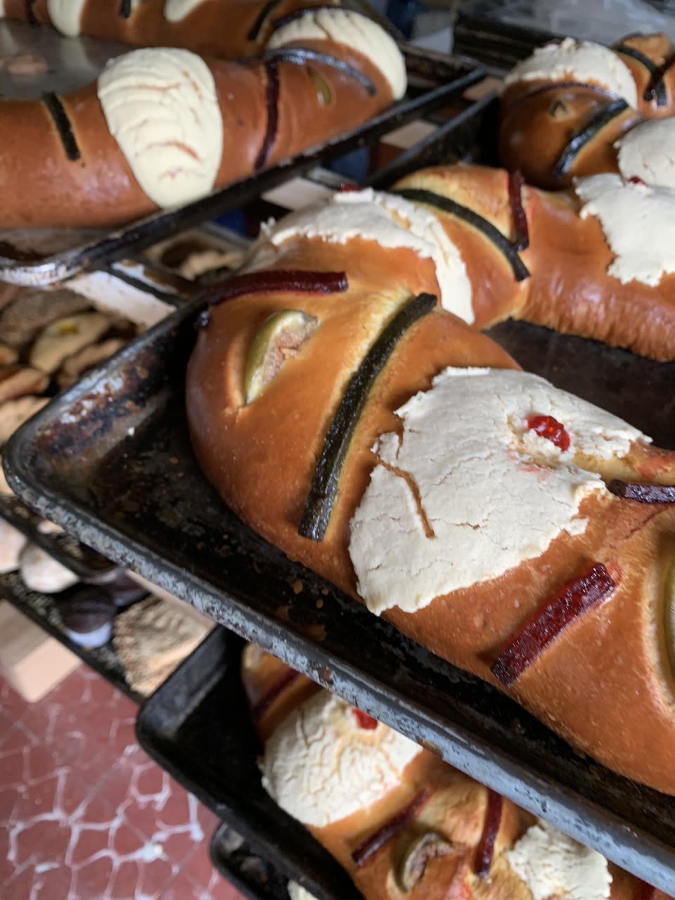Several roscas de reyes stacked on blackened steel baking trays on a bakery rack, photographed close and from slightly above. Each is a wide oval ring of glossy golden-brown enriched bread, scored and risen, decorated along the top with broad ovals of pale cream sugar paste and with strips of candied fruit in dark green and deep red laid between them; one strip carries a single bright red glacé cherry. The trays are old and scorched around the edges. More roscas are visible on the tray above and the tray below, and a patterned tiled floor shows at the bottom left. Nothing is cut, so none of the hidden figures can be seen. No people are in the picture.