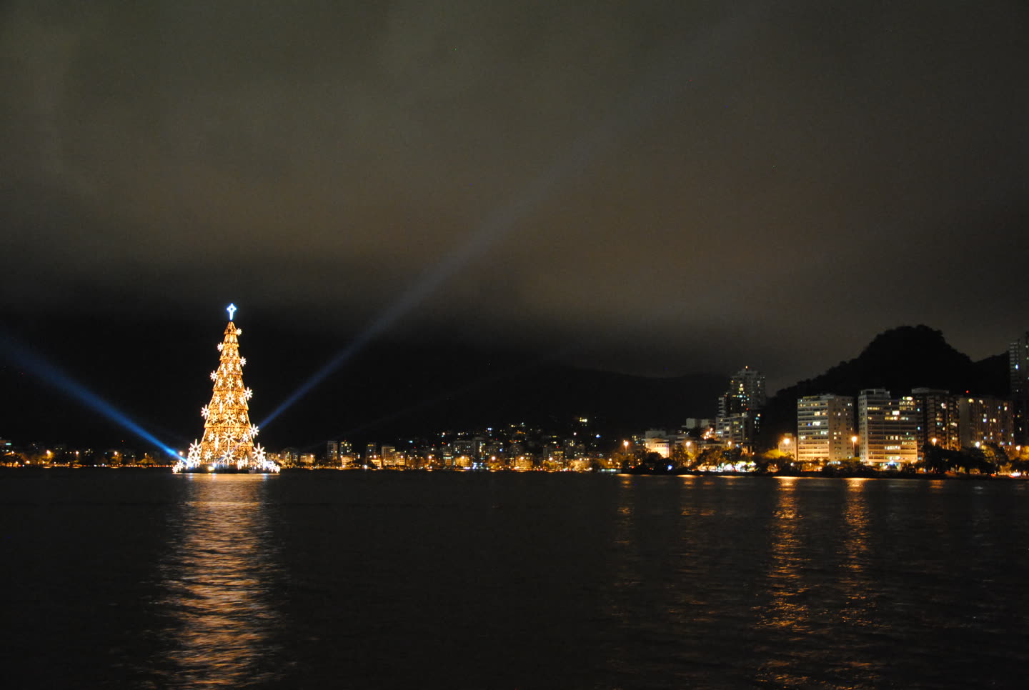 A huge illuminated Christmas tree standing on the water at night, seen from across the Lagoa Rodrigo de Freitas in Rio de Janeiro. The tree is a tall tapering steel frame outlined in warm golden lights, hung with large white star shapes and topped by a pale blue star, mounted on a dark floating platform at the left of the frame. Two searchlight beams angle up and outward from its base into a low overcast sky. Along the far shore the lit windows and street lamps of apartment blocks run from the centre of the picture to the right, and the dark bulk of a forested hill rises behind them at the right-hand edge. The lagoon fills the lower third, black and faintly rippled, carrying a long golden reflection of the tree and shorter broken reflections of the shore lights. No people are visible.