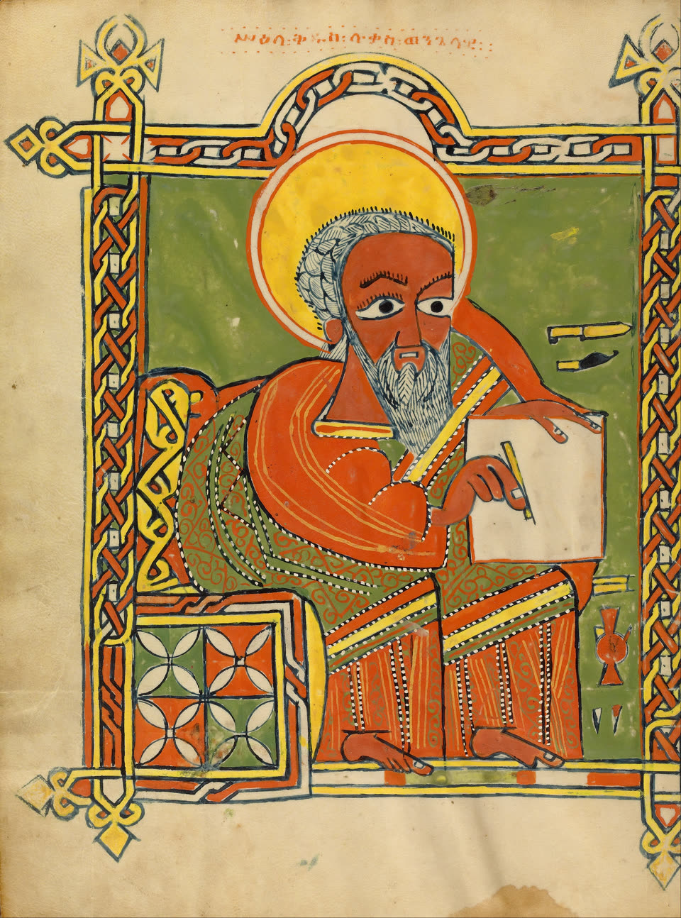 A full-page portrait of Saint Luke from an Ethiopian Gospel book, painted in flat opaque colour on parchment. The evangelist sits on a cushioned stool turned three-quarters to the viewer, his face and hands brick red, his eyes very large and dark, a curled white beard and white hair beneath an enormous yellow halo ringed in orange. He wears a vermilion cloak over a green and gold robe patterned with scrolls, steadies a blank sheet with one hand and writes on it with a reed pen in the other, and rests his bare feet on a step. The ground behind him is plain green, with a pen-case and a small knife hanging at the right. A broad band of red, yellow and white interlace frames the whole page, with cross-shaped finials at its corners, and a line of red Geʽez script runs across the top.