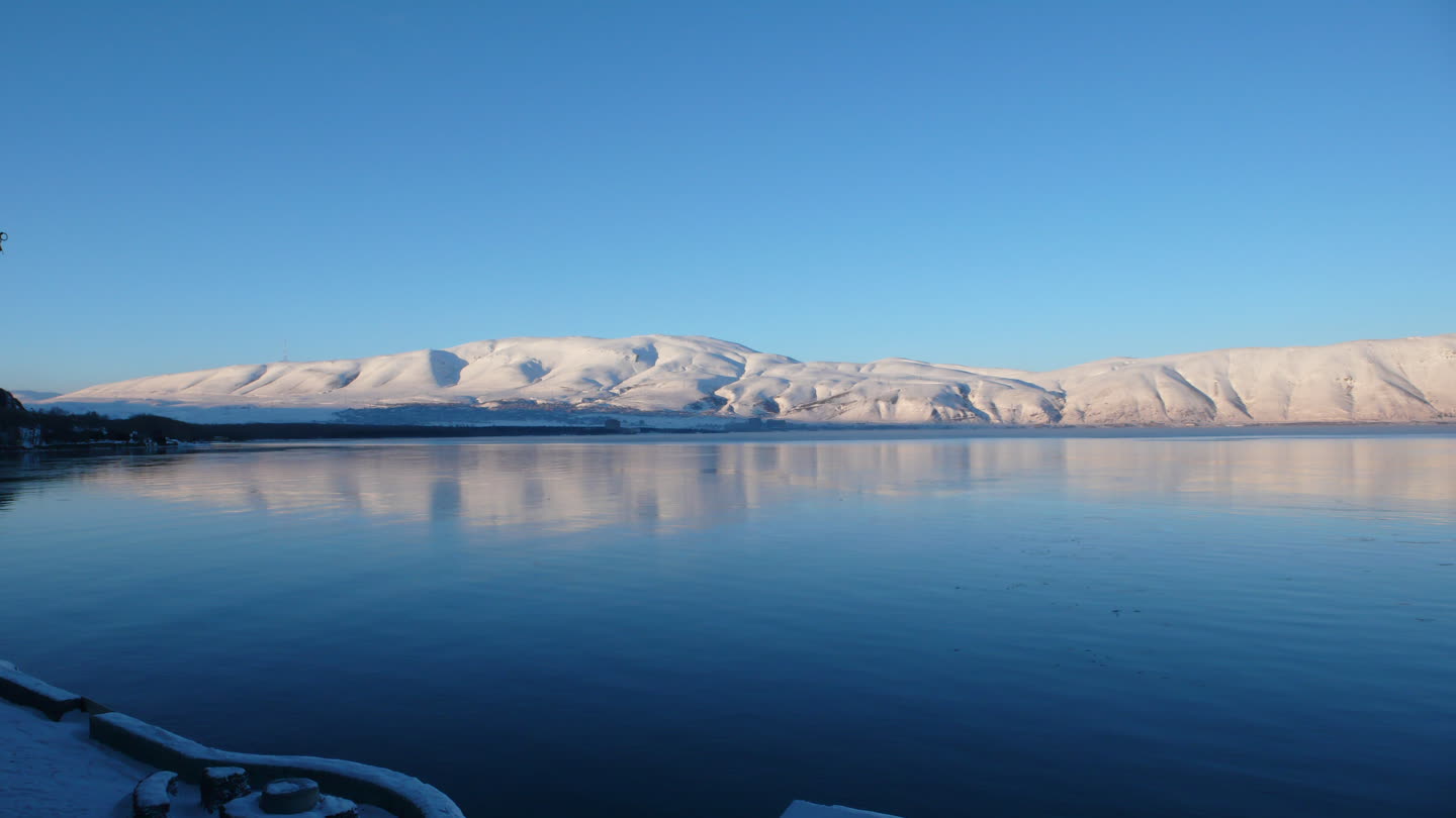 Lake Sevan in winter under a clear pale-blue sky: a long ridge of snow-covered hills along the far shore catching low sun, the white slopes mirrored in still dark-blue water, and snow lying on the stone quay in the near foreground.