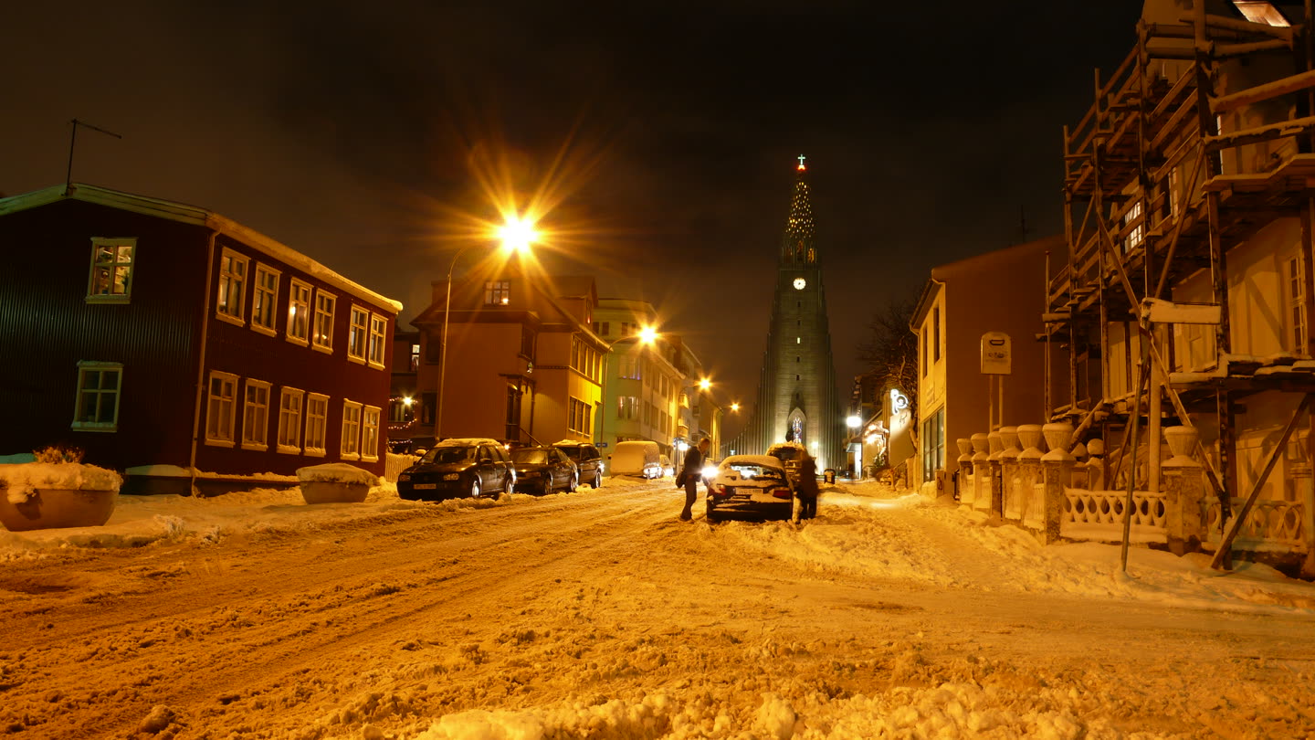 A snowy Reykjavik street at night: a slush-covered road between timber and corrugated-iron houses, cars under sodium streetlamps, two people by a car, and the tower of Hallgrimskirkja at the top of the hill strung with Christmas lights and a star.