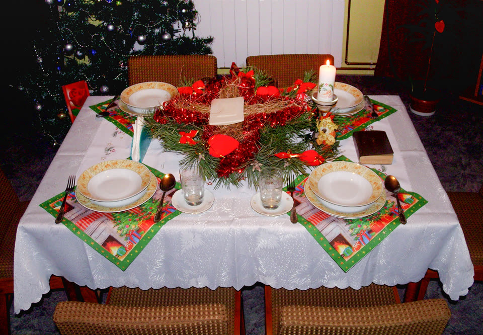 A Wigilia table laid for four and not yet eaten from: a white cloth over a square table, four places set with deep white bowls on patterned plates, forks and spoons on Christmas napkins, a tumbler at each. In the middle a ring of pine, red berries and ribbons around a loose heap of hay, and lying on the hay a folded napkin and the pale rectangles of the opłatek. A tall candle burns at the back beside a dark prayer book, and a lit Christmas tree stands in the dark behind the table.