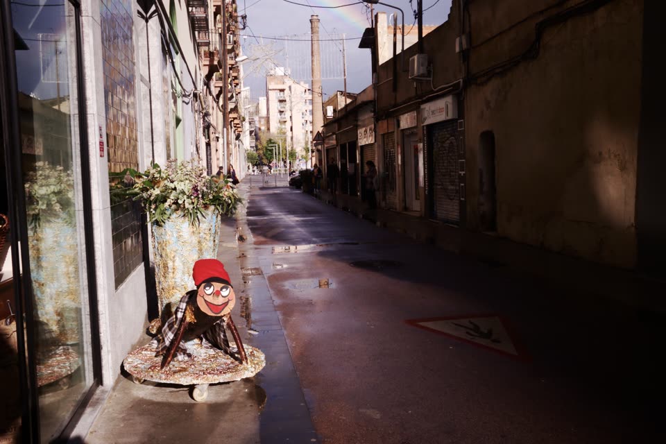 A tio standing on the pavement of a narrow Catalan street after rain, in the modern form the custom has settled into: a short log with a grinning face painted on its sawn end, round white eyes, a red barretina pulled over the top, a checked blanket across its back and two bare branches for front legs, propped on a straw mat at the foot of a shop window. The street behind it is wet and dark and runs between shuttered shopfronts towards a factory chimney, with Christmas lights strung overhead and the faint arc of a rainbow in a storm-coloured sky. Three or four people stand far down the street, too distant to make out.