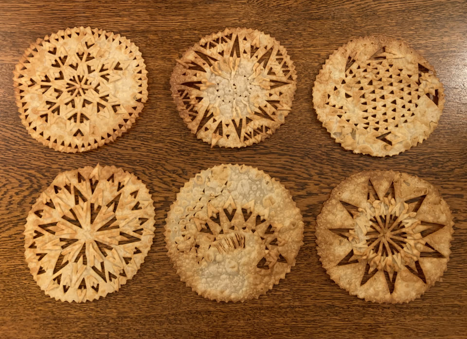 Six discs of laufabraud laid out on a dark wooden table, each cut by hand into a different fretwork pattern of triangles and stars, fried until crisp.