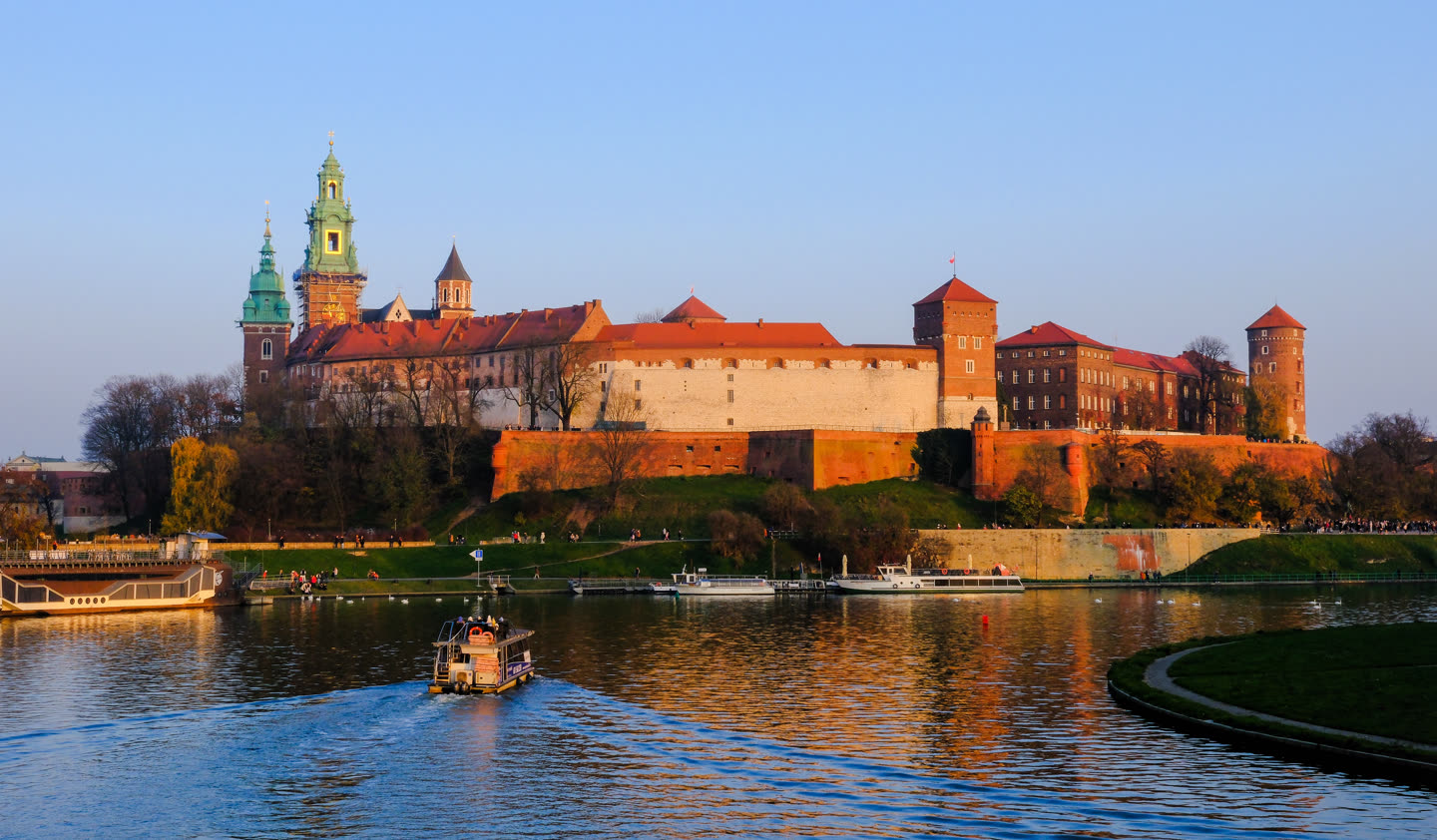 Wawel seen across the Vistula in low evening sun: the castle ridge running the width of the picture, its long red roofs and whitewashed walls above a steep brick bastion, the cathedral at the left with two green copper domes and a gilded cupola, a square brick tower with a red flag at the centre and the round Sandomierska tower at the right. Below, a grassy bank and a stone embankment with moored river boats, and a small river tram crossing the water towards the camera, leaving a wake across the reflected towers.