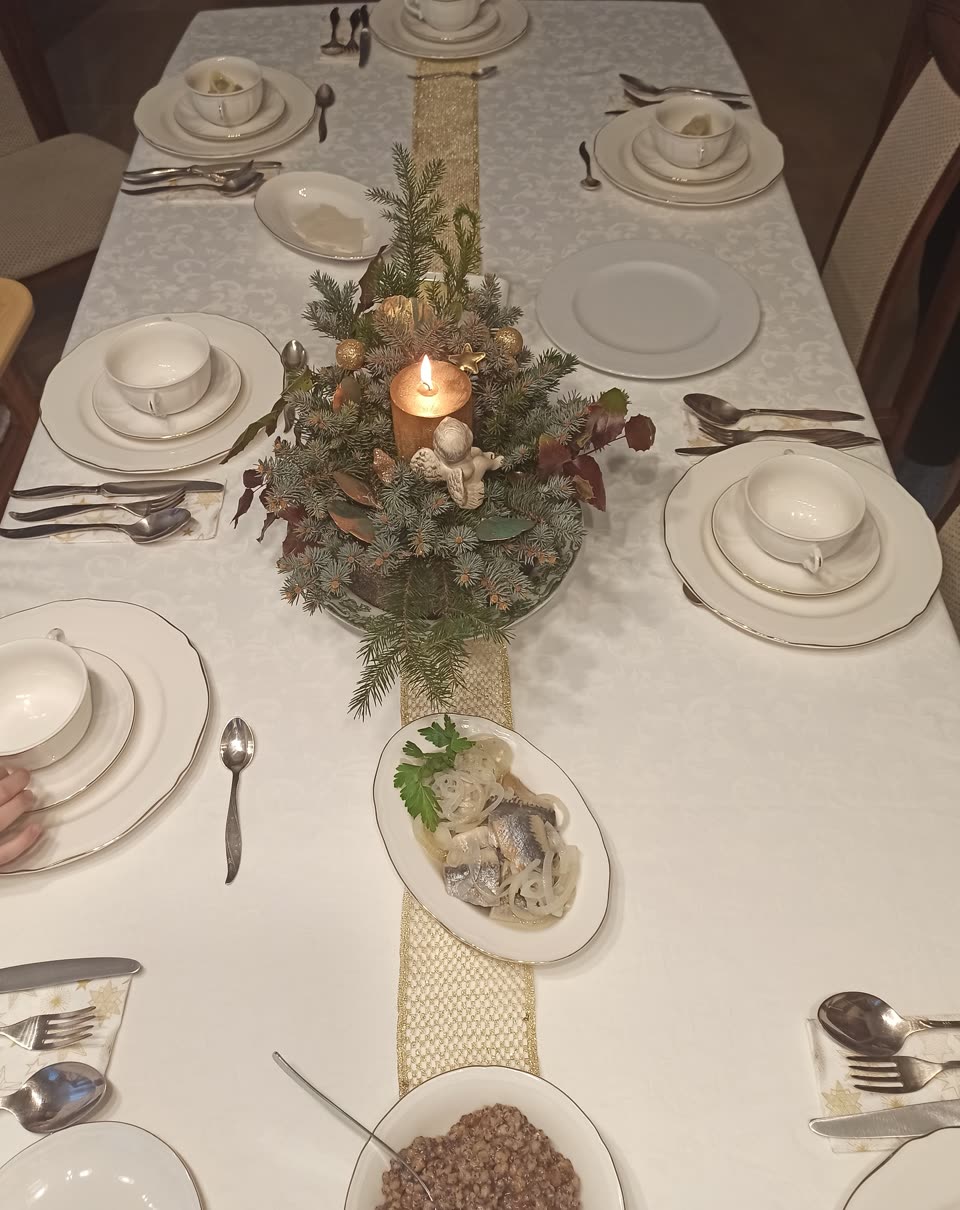 A long Wigilia table seen down its length, laid on a white damask cloth with a gold runner: seven or eight places set with gold-rimmed white china, a lidded soup cup on a plate at each, cutlery on star-printed napkins. A lit gold candle stands in a centrepiece of spruce, copper leaves and baubles with a small ceramic angel at its foot. Nearer the camera an oval dish of herring in onion rings with a sprig of parsley, and at the bottom edge a bowl of grain dark with poppy seed. A plate of pale wafer pieces stands at the far end. Somebody's fingers rest on the cloth at the left edge.